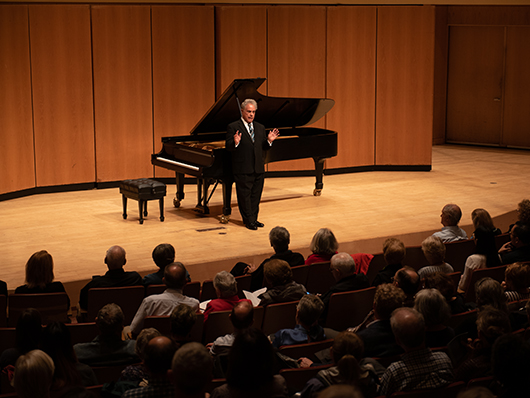 Jeffrey Siegel stands onstage in front of a grand piano, speaking to the audience in Pick-Staiger Concert Hall